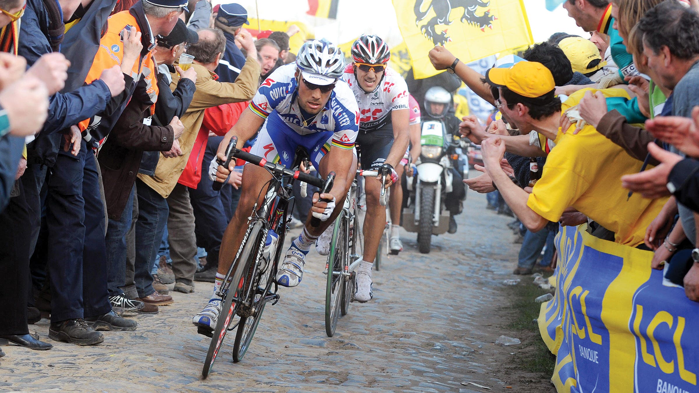 Boonen rides over a narrow cobblestone street ahead of Cancellara with cheering fans on either side