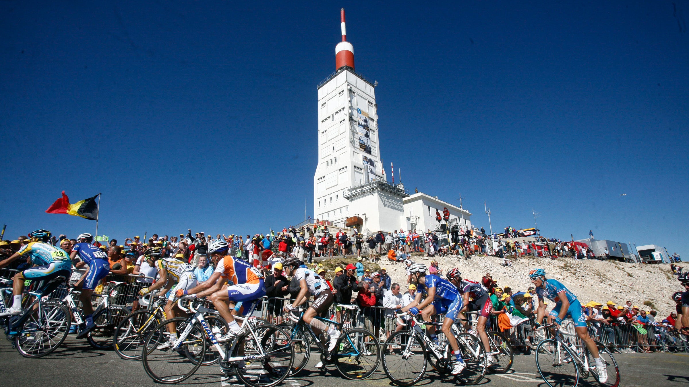 Large group of cyclists riding by a white observation tower at a mountaintop