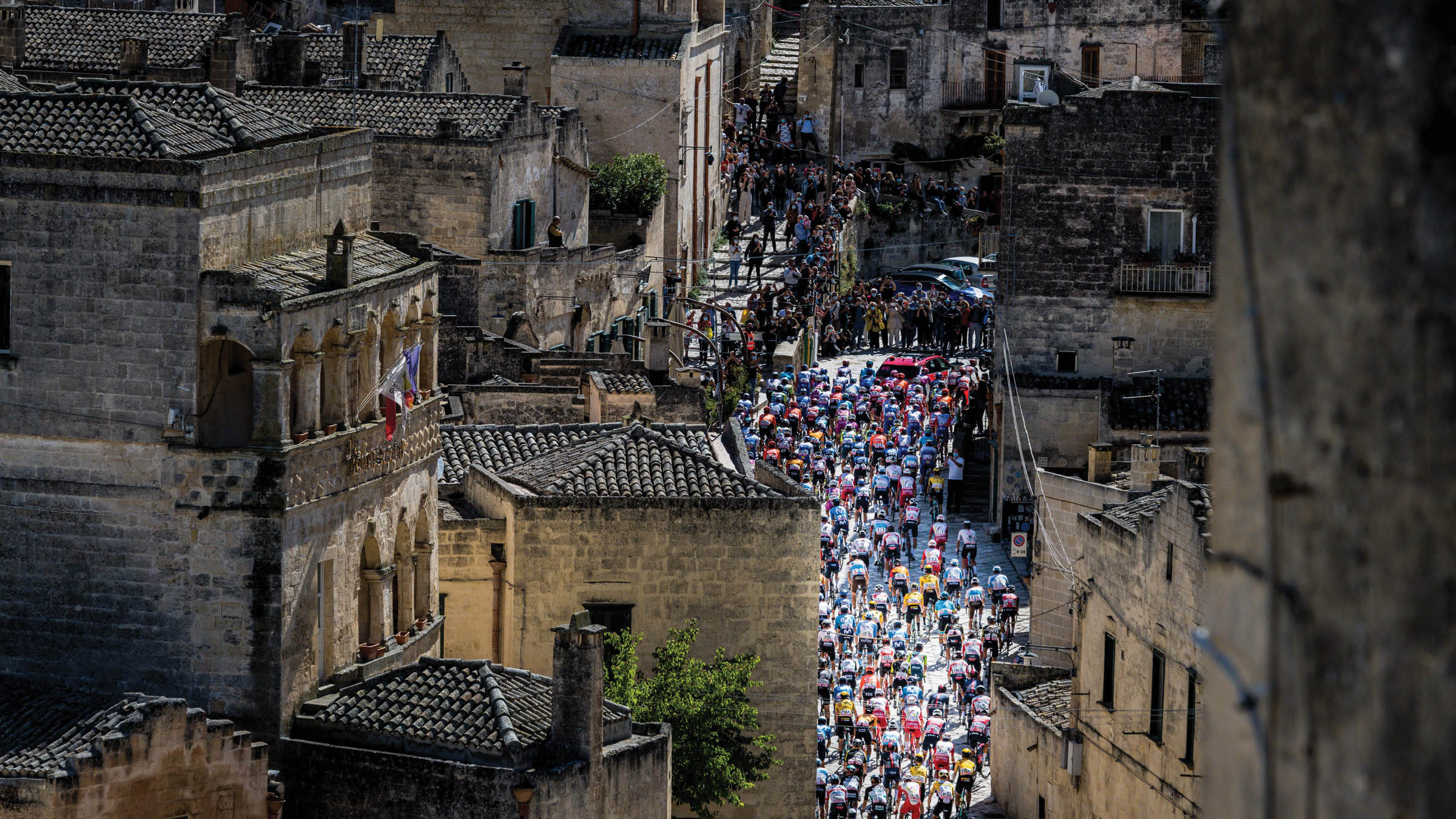 Large group of cyclists ride through tall brick buildings of Matera, Italy